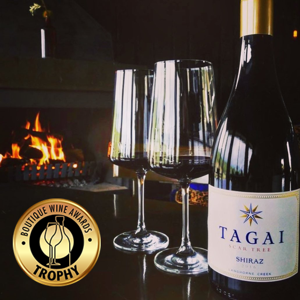 Bottle of Tagai Shiraz next to 2 glasses of wine. With a roaring fire in the background. Overlaid text and Boutique Wine Awards Trophy sticker.